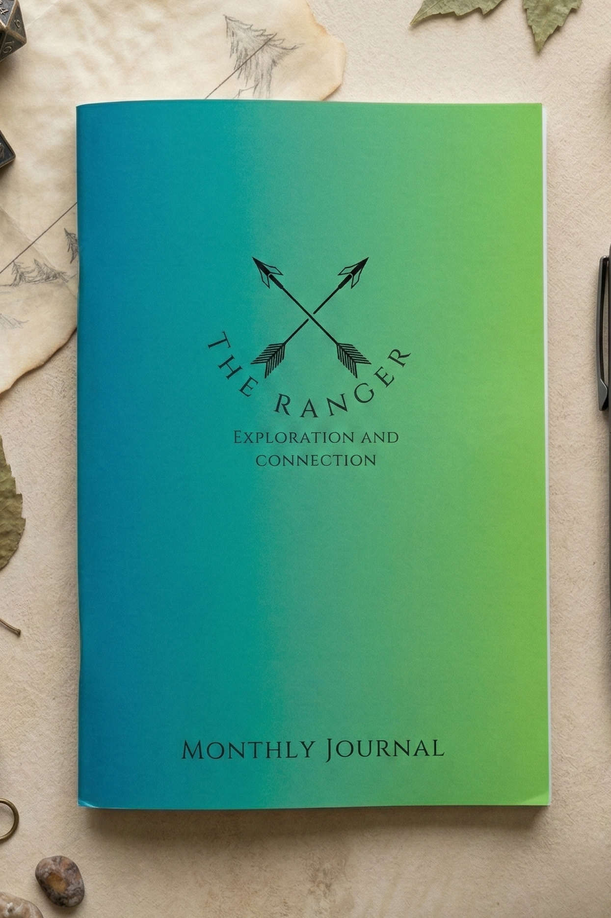 A monthly journal with a blue-to-green gradient cover, featuring the text 'THE RANGER' and 'EXPLORATION AND CONNECTION' with crossed arrows, plus 'MONTHLY JOURNAL' at the bottom. The journal is photographed flat on a textured beige surface with scattered dried leaves and small stones around it.
